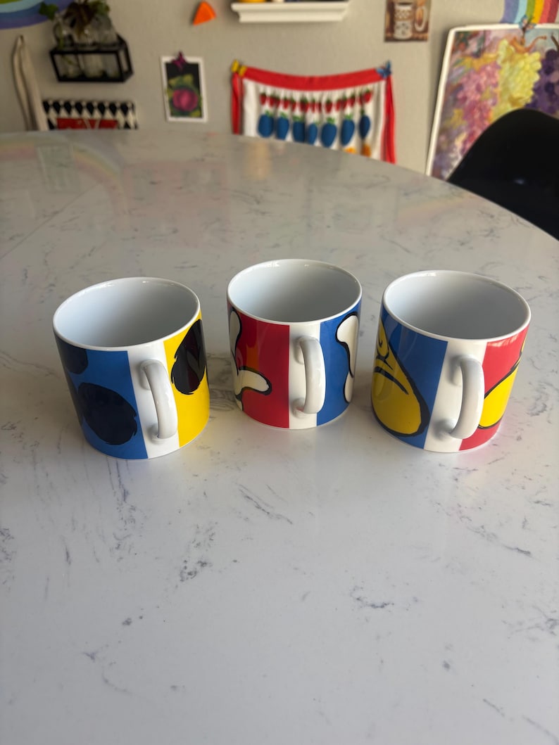 K&ouml;nnte beinhalten: Drei Keramikbecher mit farbenfrohen abstrakten Designs. Jeder Becher hat einen wei&szlig;en Henkel und ein einzigartiges Muster aus blauen, roten und gelben Formen. Die Becher stehen auf einer wei&szlig;en Oberfl&auml;che.