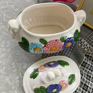 May include: White ceramic serving bowl with a floral design. The bowl has a lid with a floral design and a knob on top. The bowl is decorated with pink, blue, and yellow flowers.