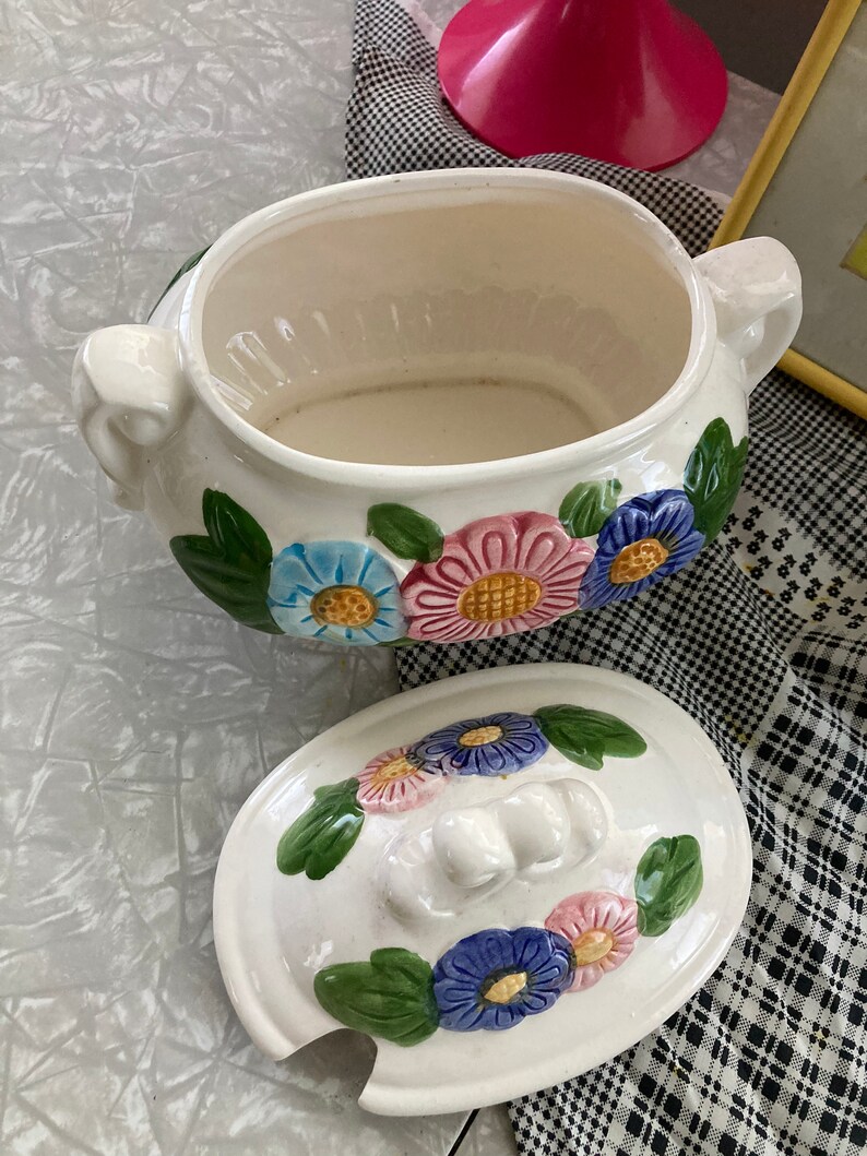 May include: White ceramic serving bowl with a floral design. The bowl has a lid with a floral design and a knob on top. The bowl is decorated with pink, blue, and yellow flowers.