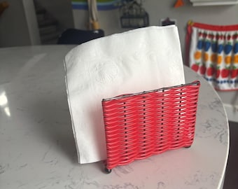 May include: A red woven napkin holder with a stack of white napkins. The napkin holder is rectangular and has a woven design. The napkins are white with a seashell pattern. The items are on a marble-like surface.