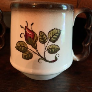 May include: A white ceramic mug with a brown rim and handle. The mug has a floral design featuring a red rose bud and green leaves.