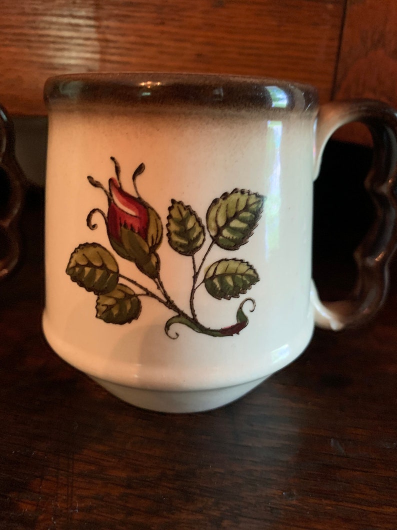 May include: A white ceramic mug with a brown rim and handle. The mug has a floral design featuring a red rose bud and green leaves.