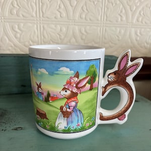 May include: White ceramic mug with a whimsical Easter bunny design. The mug features a wrap-around illustration of bunnies in a pastoral setting, with a bunny-shaped handle. The bunny handle is brown with pink inner ears.