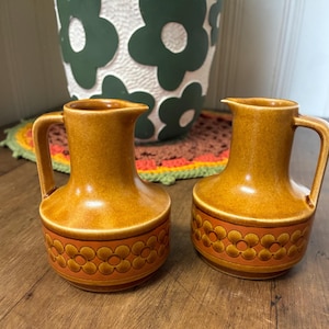 May include: Two vintage ceramic pitchers with a warm, earthy color palette. The pitchers have a rounded base with a band of orange floral designs. Each pitcher has a handle and a spout. The background includes a decorative pot and a colorful coaster.