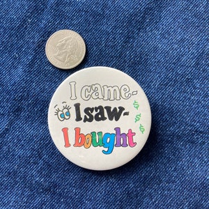 May include: A white button with the text "I came, I saw, I bought" in rainbow colors. The button has a cartoon eye with eyelashes on the left side.
