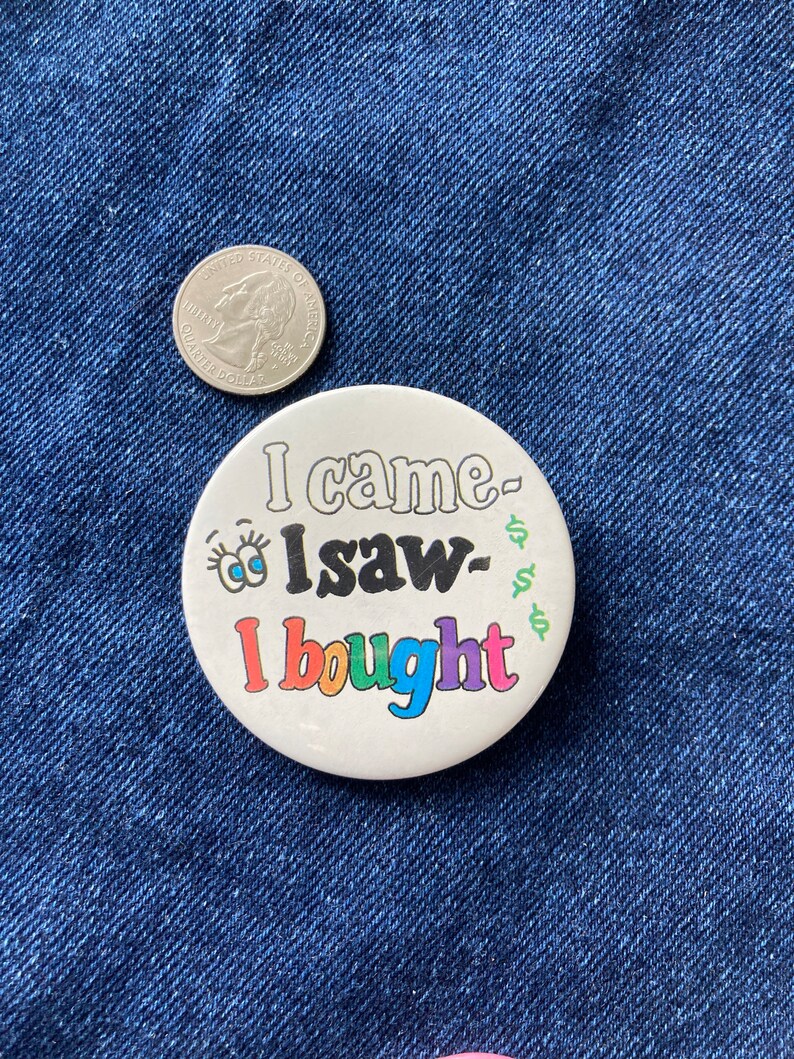 May include: A white button with the text "I came, I saw, I bought" in rainbow colors. The button has a cartoon eye with eyelashes on the left side.