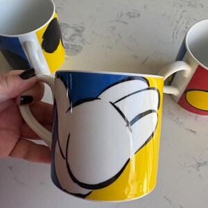 K&ouml;nnte beinhalten: Drei wei&szlig;e Keramikbecher mit farbenfrohen Designs. Ein Becher zeigt eine wei&szlig;e Hand auf gelbem und blauem Hintergrund. Die anderen Becher haben &auml;hnliche Designs mit verschiedenen Farben. Jeder Becher hat einen wei&szlig;en Henkel.