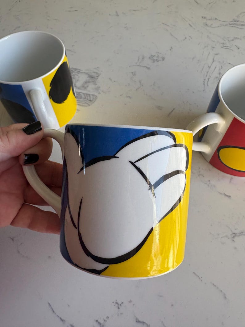 K&ouml;nnte beinhalten: Drei wei&szlig;e Keramikbecher mit farbenfrohen Designs. Ein Becher zeigt eine wei&szlig;e Hand auf gelbem und blauem Hintergrund. Die anderen Becher haben &auml;hnliche Designs mit verschiedenen Farben. Jeder Becher hat einen wei&szlig;en Henkel.