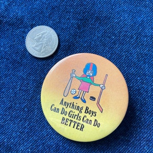 May include: A yellow button with a cartoon illustration of a girl playing sports. The text on the button reads "Anything Boys Can Do Girls Can Do BETTER."