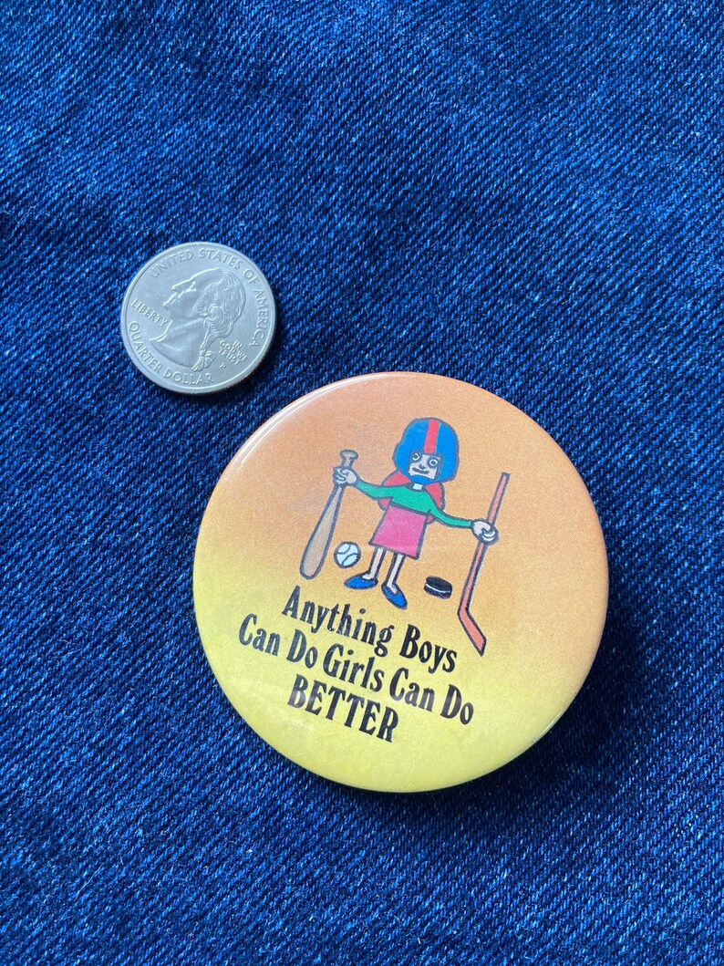 May include: A yellow button with a cartoon illustration of a girl playing sports. The text on the button reads "Anything Boys Can Do Girls Can Do BETTER."