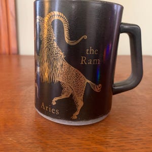 May include: Black ceramic mug with a gold illustration of the Aries zodiac sign, a ram, with the text "the Ram" and "Aries".