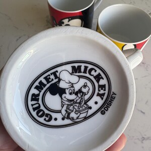 K&ouml;nnte beinhalten: Drei wei&szlig;e Keramikbecher mit farbenfrohen Mickey Mouse-Designs. Ein Becher zeigt Mickys Gesicht in Rot und Schwarz, ein anderer in Gelb und Schwarz, und der dritte hat ein "Gourmet Mickey"-Logo.