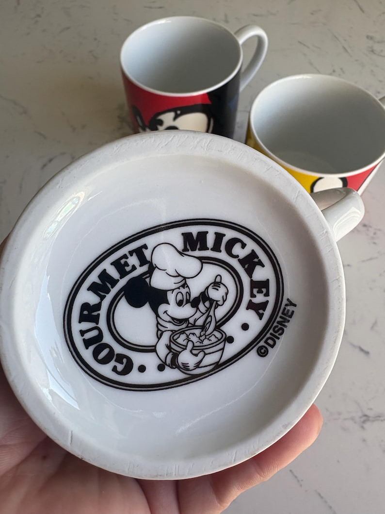 K&ouml;nnte beinhalten: Drei wei&szlig;e Keramikbecher mit farbenfrohen Mickey Mouse-Designs. Ein Becher zeigt Mickys Gesicht in Rot und Schwarz, ein anderer in Gelb und Schwarz, und der dritte hat ein "Gourmet Mickey"-Logo.