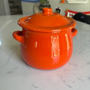 May include: A bright orange ceramic pot with a matching lid and two side handles. The pot features a smooth, glossy finish and a rounded body shape. The lid has a small central knob for lifting, and the entire piece is a solid, vibrant orange color.