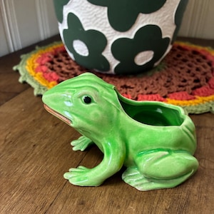 May include: A ceramic frog planter in a bright green glaze. The frog has a wide open mouth and a hollowed-out back for planting. The planter is on a wooden surface, with a decorative doily and a flower-patterned pot in the background.