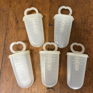 May include: Five clear, plastic Tupperware ice pop molds with handles. Each mold has a rounded bottom and a handle at the top. The text "Tupperware" and "Made in U.S.A." is visible on each mold.