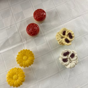May include: Six ceramic flower-shaped drawer knobs in red, yellow, and white. The knobs feature a rose, pansy, and daisy design.