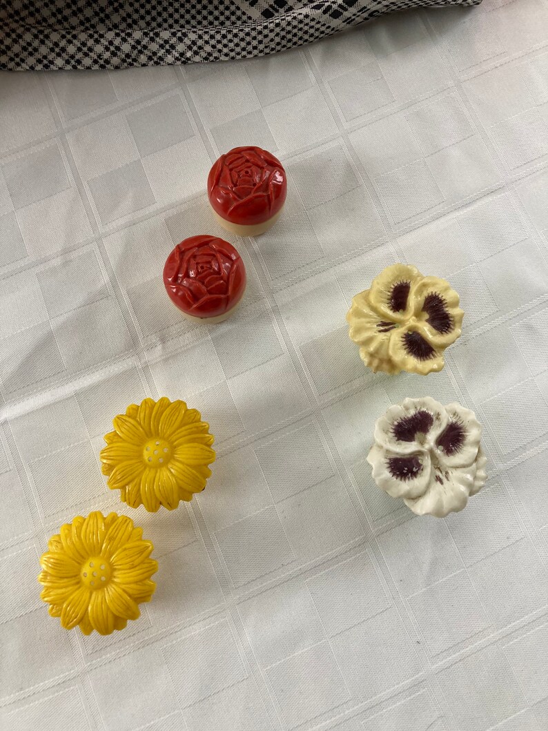 May include: Six ceramic flower-shaped drawer knobs in red, yellow, and white. The knobs feature a rose, pansy, and daisy design.