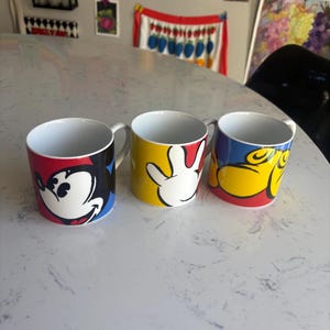 K&ouml;nnte beinhalten: Drei Keramikbecher mit Mickey-Mouse-Designs. Ein Becher zeigt Mickys Gesicht, ein anderer seine Hand und der dritte sein Gesicht in einer anderen Pose. Jeder Becher hat ein wei&szlig;es Innere und einen Henkel.