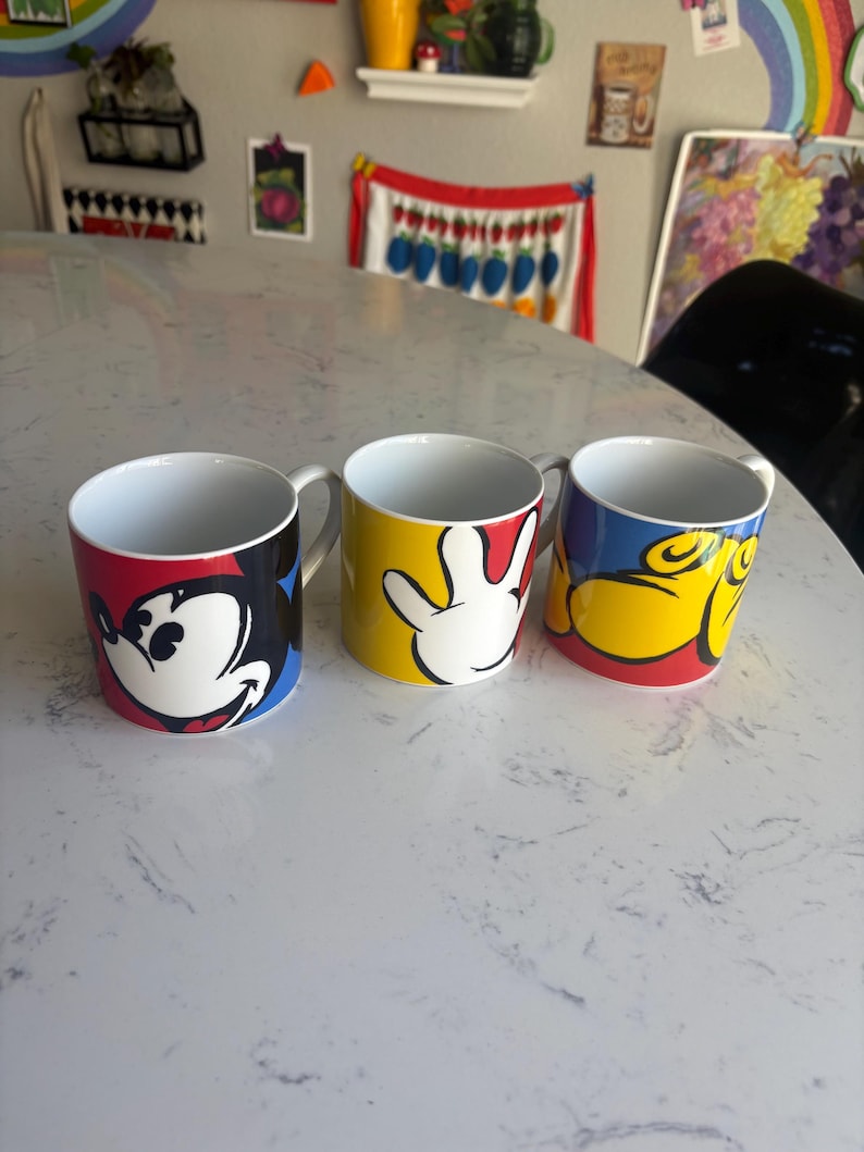 K&ouml;nnte beinhalten: Drei Keramikbecher mit Mickey-Mouse-Designs. Ein Becher zeigt Mickys Gesicht, ein anderer seine Hand und der dritte sein Gesicht in einer anderen Pose. Jeder Becher hat ein wei&szlig;es Innere und einen Henkel.