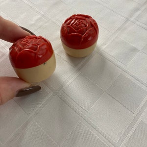 May include: Two small, round, red and white containers with a rose design on the top. The containers are made of plastic.