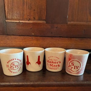 May include: Four off-white shot glasses with red graphics and text. Two glasses feature the Maker's Mark logo, one with a starburst design, and one with two red drips. The glasses are on a dark wooden surface.