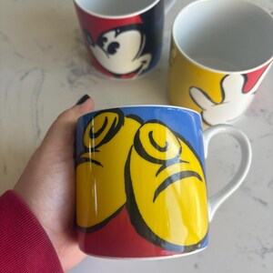 K&ouml;nnte beinhalten: Drei wei&szlig;e Keramikbecher mit Cartoon-Charakter-Designs. Ein Becher zeigt Mickey Mouses Gesicht, ein anderer seine Hand, und der dritte Becher hat ein Design von Mickey Mouses Gesicht und H&auml;nden. Die Becher haben ein wei&szlig;es Innere und einen Henkel.