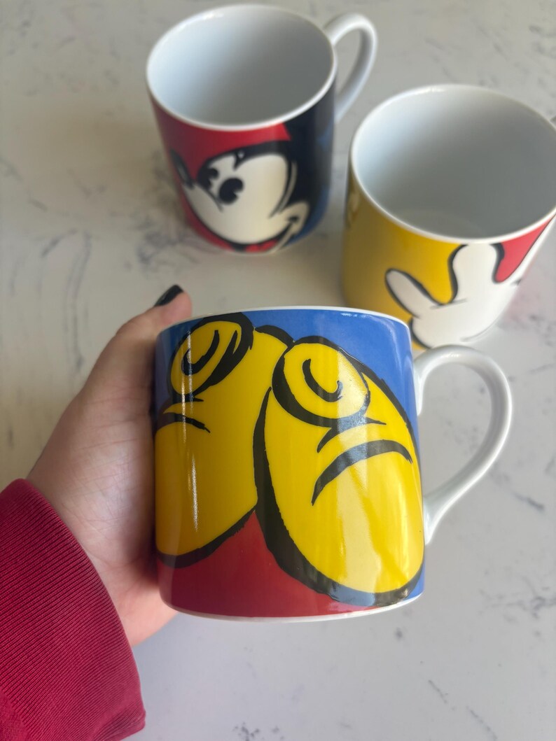 K&ouml;nnte beinhalten: Drei wei&szlig;e Keramikbecher mit Cartoon-Charakter-Designs. Ein Becher zeigt Mickey Mouses Gesicht, ein anderer seine Hand, und der dritte Becher hat ein Design von Mickey Mouses Gesicht und H&auml;nden. Die Becher haben ein wei&szlig;es Innere und einen Henkel.
