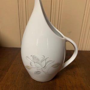 May include: White ceramic pitcher with a single gray rose design. The pitcher has a unique, curved spout and a handle.