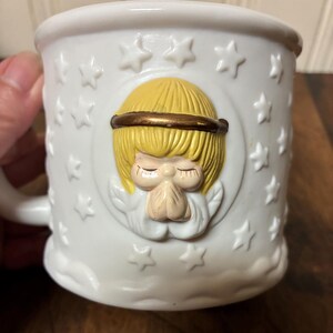 May include: White ceramic mug with a raised relief design of an angel with folded hands, a gold halo, and blonde hair. The mug is decorated with embossed stars and a cloud-like base.