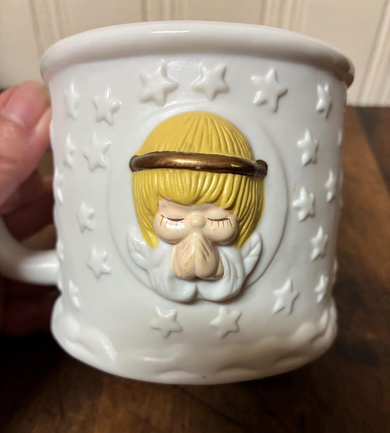 May include: White ceramic mug with a raised relief design of an angel with folded hands, a gold halo, and blonde hair. The mug is decorated with embossed stars and a cloud-like base.