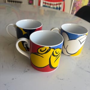 K&ouml;nnte beinhalten: Drei Keramikbecher mit wei&szlig;en Innenr&auml;umen und Griffen. Die Becher haben farbenfrohe Designs mit roten, gelben und blauen Akzenten. Ein Becher hat ein Design einer Cartoon-Hand, ein anderer ein Design eines Cartoon-Gesichts und der dritte ein Sterndesign.