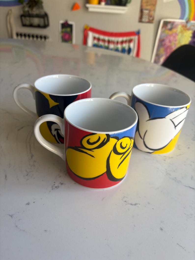 K&ouml;nnte beinhalten: Drei Keramikbecher mit wei&szlig;en Innenr&auml;umen und Griffen. Die Becher haben farbenfrohe Designs mit roten, gelben und blauen Akzenten. Ein Becher hat ein Design einer Cartoon-Hand, ein anderer ein Design eines Cartoon-Gesichts und der dritte ein Sterndesign.