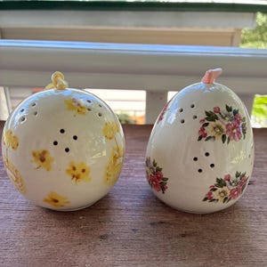 May include: Two ceramic salt and pepper shakers. One is round with yellow floral designs and a yellow top. The other is egg-shaped with pink and yellow floral designs and a pink top. Both have several small holes.