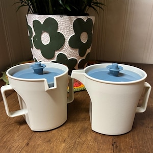 May include: Two off-white plastic pitchers with angular handles and blue circular lids featuring a central knob. Each pitcher has a small pouring spout on the side. The design is uniform, with a smooth finish and a structured, geometric shape.