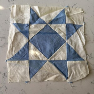May include: A handmade quilt block featuring a blue and white star design. The block is made from two different fabrics, one white and one blue, and is sewn together in a traditional patchwork style.