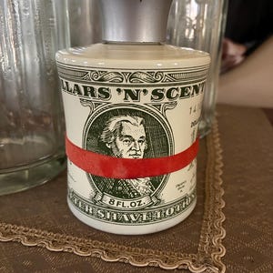 May include: A vintage shaving lotion bottle with a silver cap and a cream-colored body. The bottle features a design resembling a dollar bill, with the text "DOLLARS 'N' SCENT" and "AFTER SHAVE LOTION". A red band encircles the bottle.