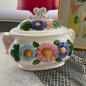 May include: White ceramic sugar bowl with a floral design. The bowl has a lid with a decorative knob and features three large flowers in shades of blue, pink, and yellow.