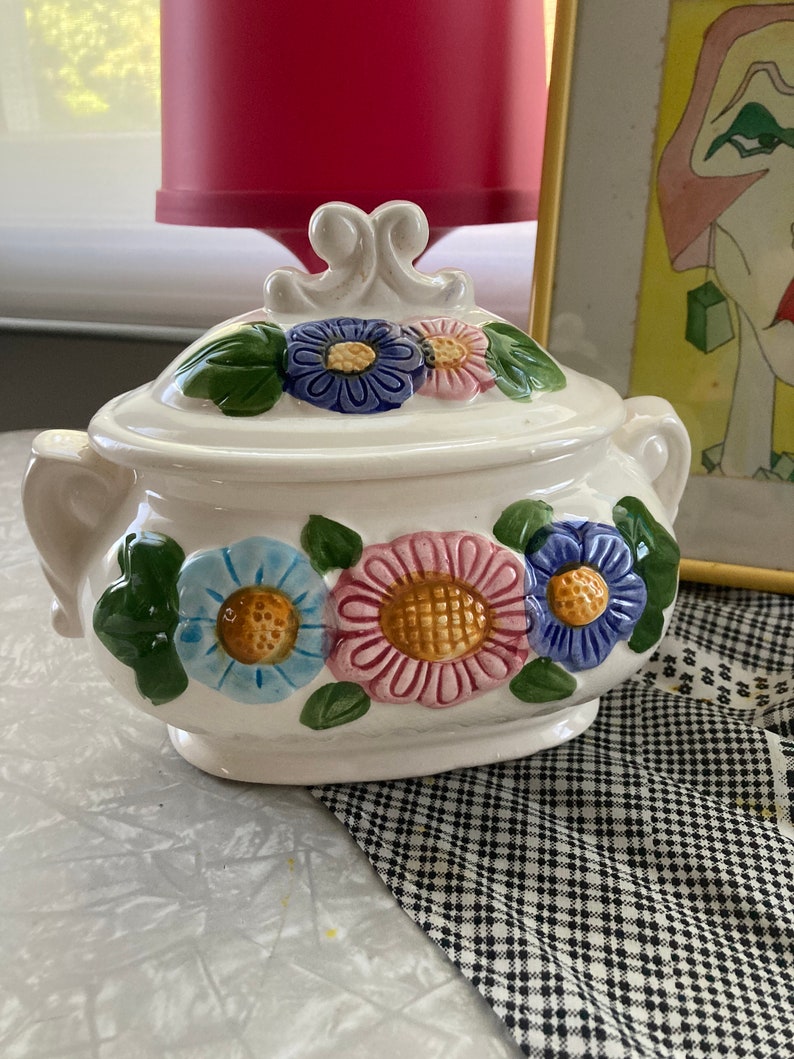 May include: White ceramic sugar bowl with a floral design. The bowl has a lid with a decorative knob and features three large flowers in shades of blue, pink, and yellow.