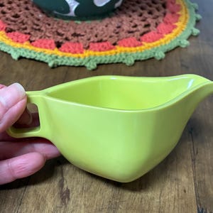 May include: A bright lime green gravy boat with a handle. The gravy boat has a spout for easy pouring. The handle is square. The gravy boat is sitting on a wooden surface.