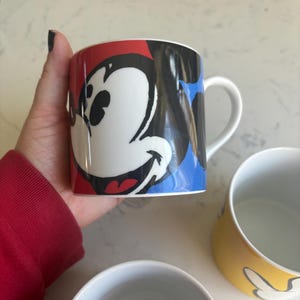 K&ouml;nnte beinhalten: Eine wei&szlig;e Keramik-Tasse mit einem farbenfrohen Mickey Mouse-Design. Die Tasse hat ein rot, schwarz und blaues Farbschema mit einem wei&szlig;en Henkel. Weitere Tassen sind im Hintergrund zu sehen, eine gelbe mit Mickey Mouse-Design und eine weitere wei&szlig;e.