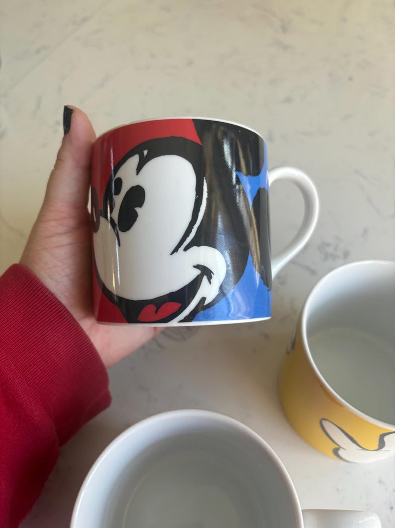 K&ouml;nnte beinhalten: Eine wei&szlig;e Keramik-Tasse mit einem farbenfrohen Mickey Mouse-Design. Die Tasse hat ein rot, schwarz und blaues Farbschema mit einem wei&szlig;en Henkel. Weitere Tassen sind im Hintergrund zu sehen, eine gelbe mit Mickey Mouse-Design und eine weitere wei&szlig;e.