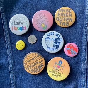 May include: A collection of nine colorful button pins with various sayings and images. The pins include: "I came, I saw, I bought", "Happy Birthday this bud's for you", "Have you hugged your dad today?", "Habe einen guten Tag", "Je t'aime", "Courtesy is contagious start an epidemic", "Anything boys can do girls can do better", a smiley face, and a coin.