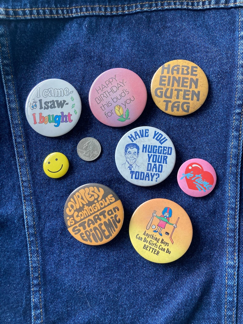 May include: A collection of nine colorful button pins with various sayings and images. The pins include: "I came, I saw, I bought", "Happy Birthday this bud's for you", "Have you hugged your dad today?", "Habe einen guten Tag", "Je t'aime", "Courtesy is contagious start an epidemic", "Anything boys can do girls can do better", a smiley face, and a coin.