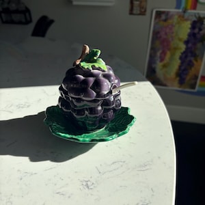 May include: A purple ceramic grape cluster honey pot with a green leaf on top and a small white spoon. The honey pot is sitting on a green ceramic leaf-shaped plate.