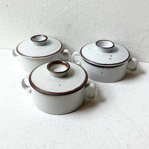 May include: Three round ceramic bowls with matching lids and side handles. Each bowl features a speckled off-white glaze with a dark brown rim around the lid and the top edge of the bowl. The design is consistent across all three pieces.