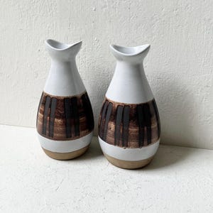 May include: Two ceramic vases with a white glazed upper section and a tapered neck. The lower body features a band of dark brown vertical stripes over a textured, light brown base. Each vase has a curved, asymmetrical rim and a rounded silhouette.