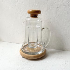 May include: A clear glass pitcher with a wooden lid and base. The pitcher has a handle and a cylindrical glass insert. The wooden base has small feet. The background is a white wall.
