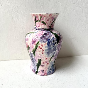 May include: A hand-painted ceramic vase with a floral design. The vase features a bulbous body and a flared rim, adorned with pink, purple, and blue flowers and green leaves. The background is a textured white.