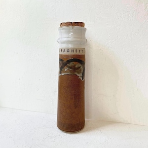 May include: A tall, brown ceramic spaghetti jar with a white top and a cork stopper. The jar has a decorative band with brown and black accents and the word "SPAGHETTI" printed on the front. The jar is cylindrical and has a textured surface.